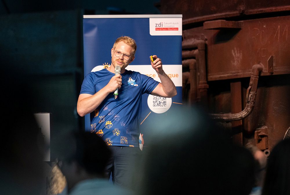 Ein Mann in einem blauen Hemd spricht bei einer Veranstaltung in einer Halle in ein Mikrofon und deutet dabei auf seine Brust, während im Hintergrund ein Werbebanner zu sehen ist.