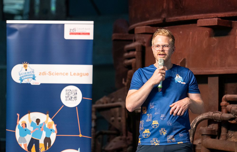 Auf der Bühne steht ein Mann in einem blauen Hemd und spricht neben einem Banner der „zdi-Science League“ in ein Mikrofon.