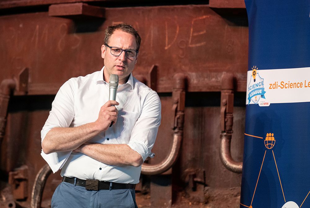 Ein Mann mit Brille und weißem Hemd spricht neben einem blauen Schild mit dem Logo der zdi-Science League in ein Mikrofon.