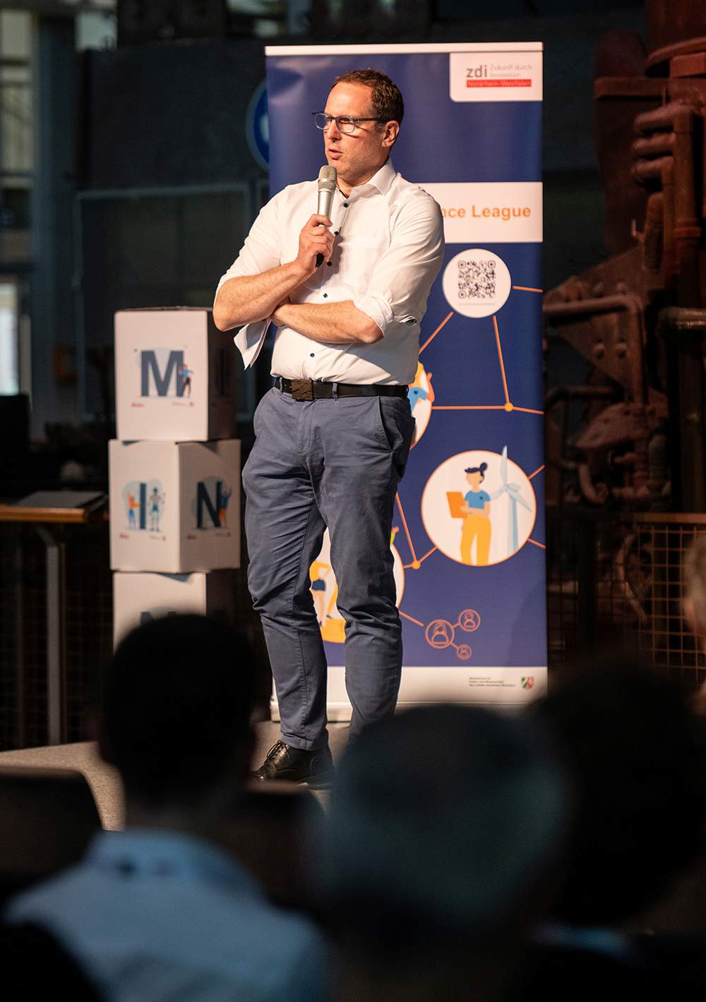 Ein Mann trägt ein weißes Hemd und eine blaue Hose und spricht auf der Bühne vor einem Banner und dem Publikum in ein Mikrofon.