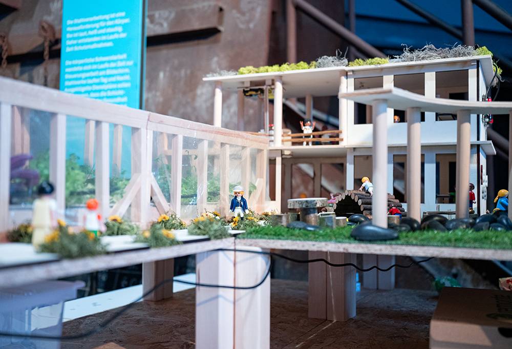 Das Bild zeigt ein detailliertes Architekturmodell eines mehrstöckigen Gebäudes mit Laufstegen, Begrünung, Minifiguren und Möbeln.