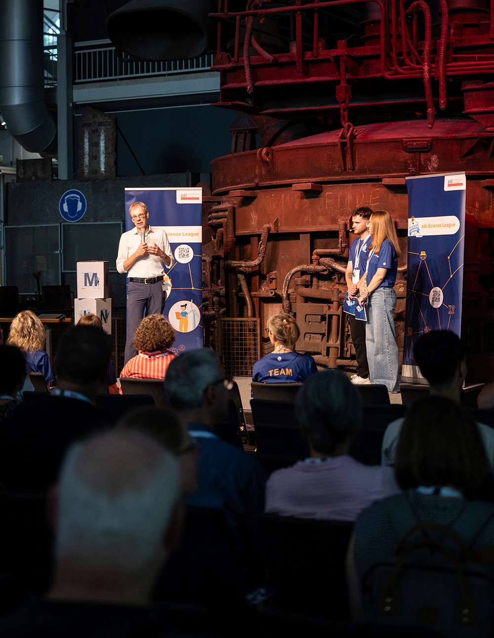 Ein Mann hält auf der Bühne eine Rede vor einem Publikum. Zwei Frauen in blauen Hemden stehen neben ihm vor Industriemaschinen in einem Innenraum.