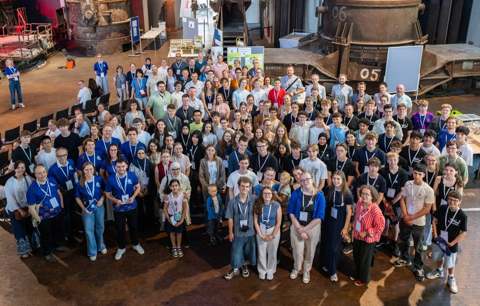 Eine große Gruppe von Menschen befindet sich bei einer Veranstaltung in einem Raum und posiert für ein Foto. Viele tragen Namensschilder und stehen vor Industriemaschinen.