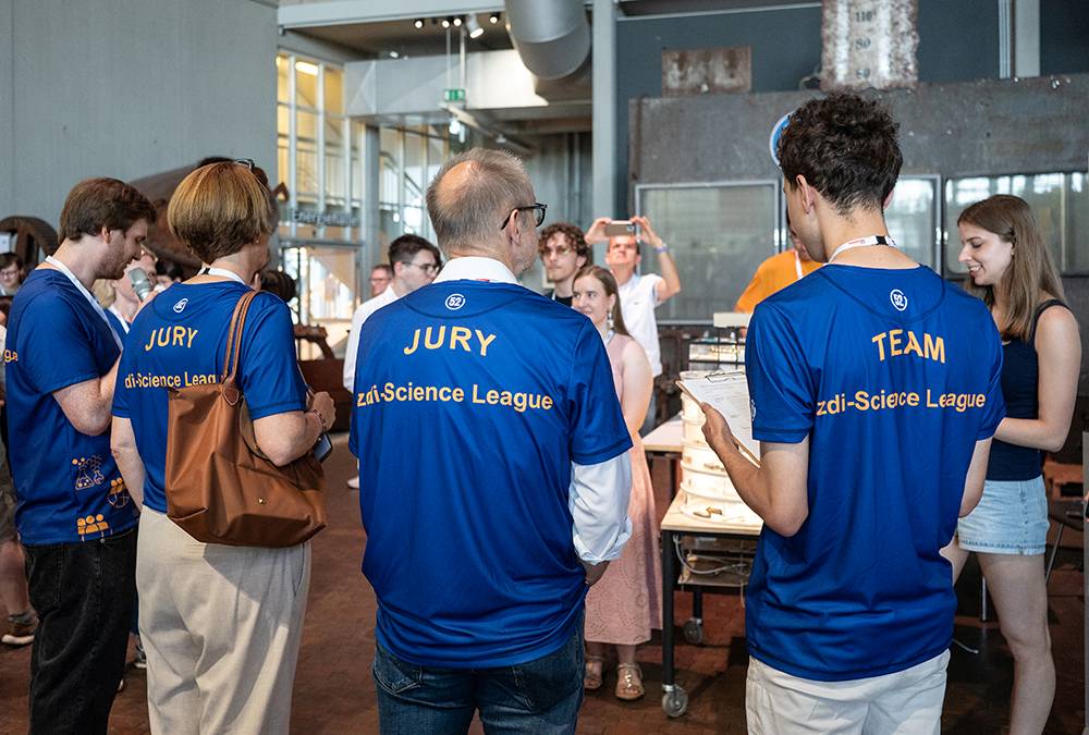 Personen in blauen T-Shirts mit den Aufschriften „JURY“ und „TEAM“ stehen bei einer Veranstaltung in einem Raum und blicken auf Tische, an denen im Hintergrund andere Teilnehmer sitzen.