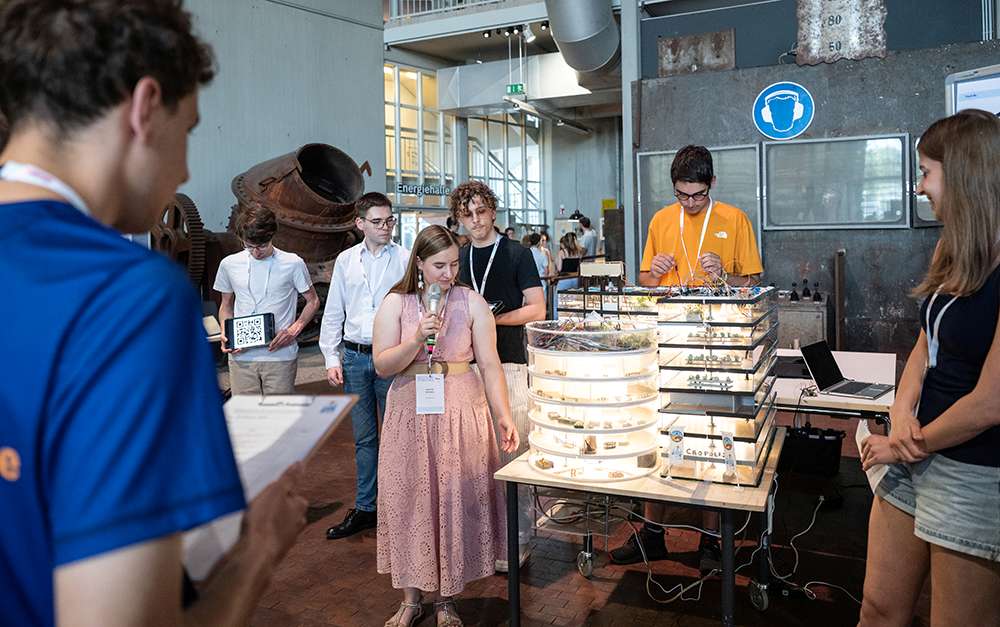 Junge Menschen versammeln sich um ein beleuchtetes Elektronikdisplay. Ein Mädchen hält ein Mikrofon in der Hand, während andere in einem Industrieraum zuschauen und sich Notizen machen.