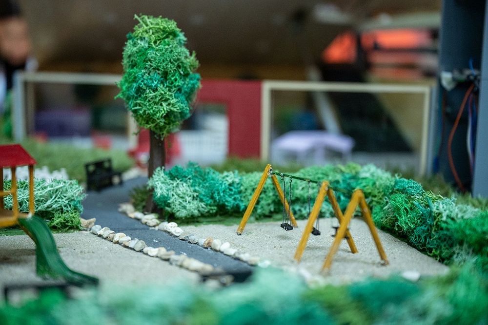Das Bild zeigt ein Miniatur-Parkmodell mit einer Schaukel, einem Baum, einer Rutsche, einer Bank und von Büschen gesäumten Wegen mit kleinen Steinen.
