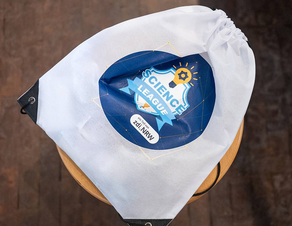 Auf einem Holzhocker, der auf einem braunen Boden steht, liegt ein weißer Beutel mit Kordelzug und einem blauen „Science League“-Logo.