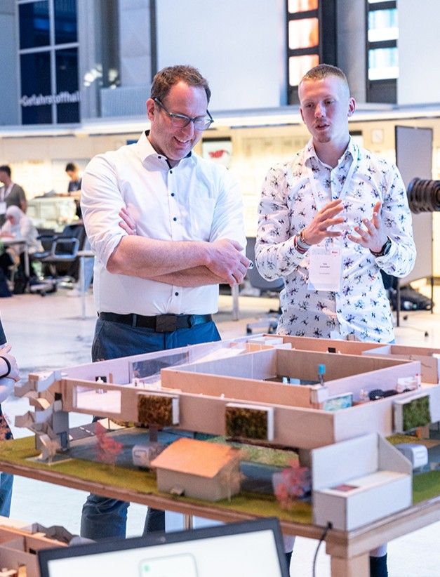 Zwei Männer stehen in einem modernen, gut beleuchteten Arbeitsbereich neben einem Tisch, auf dem ein detailliertes Architekturmodell steht, und besprechen dessen Merkmale.