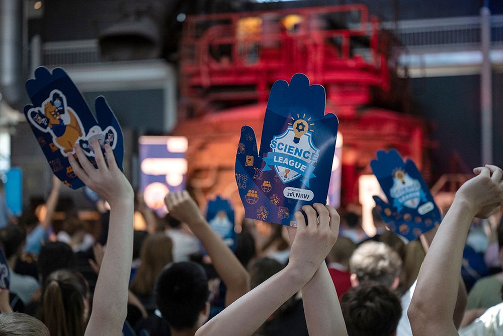 Die Besucher einer Veranstaltung in einer Halle halten Schaumstoffhände mit „Science League“-Logos in die Höhe. Im Hintergrund ist eine große rote Konstruktion zu sehen.