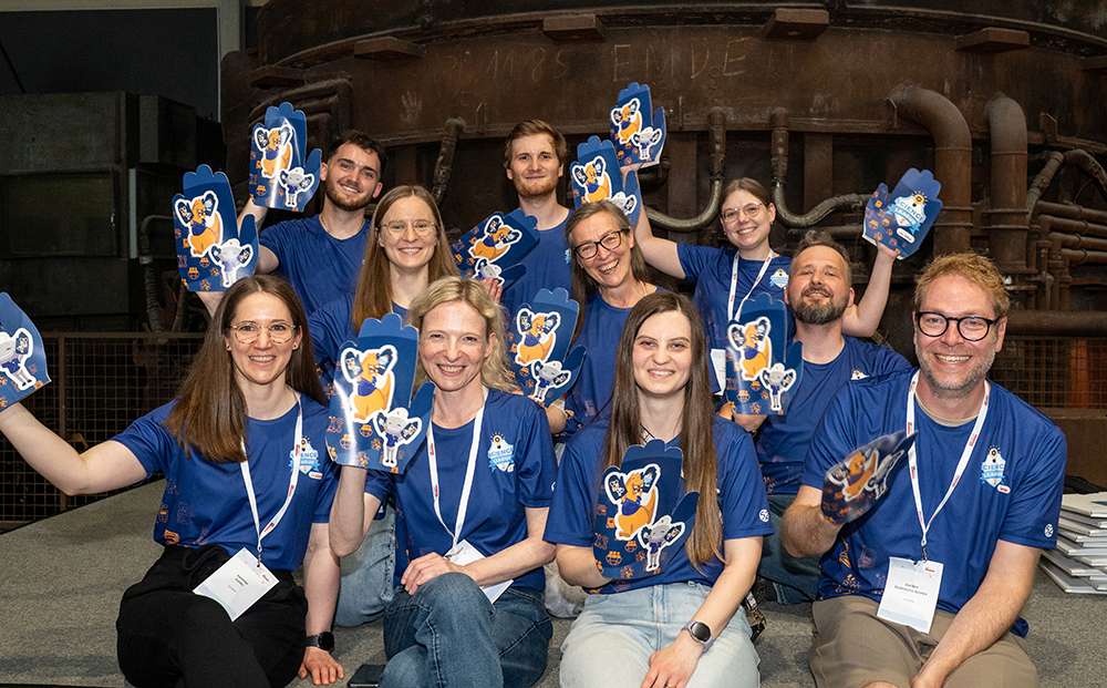 Menschen in einheitlichen blauen T-Shirts sitzen und knien gemeinsam da, lächeln und halten große blaue Schaumstoffhände mit der Abbildung einer Zeichentrickfigur in den Händen.