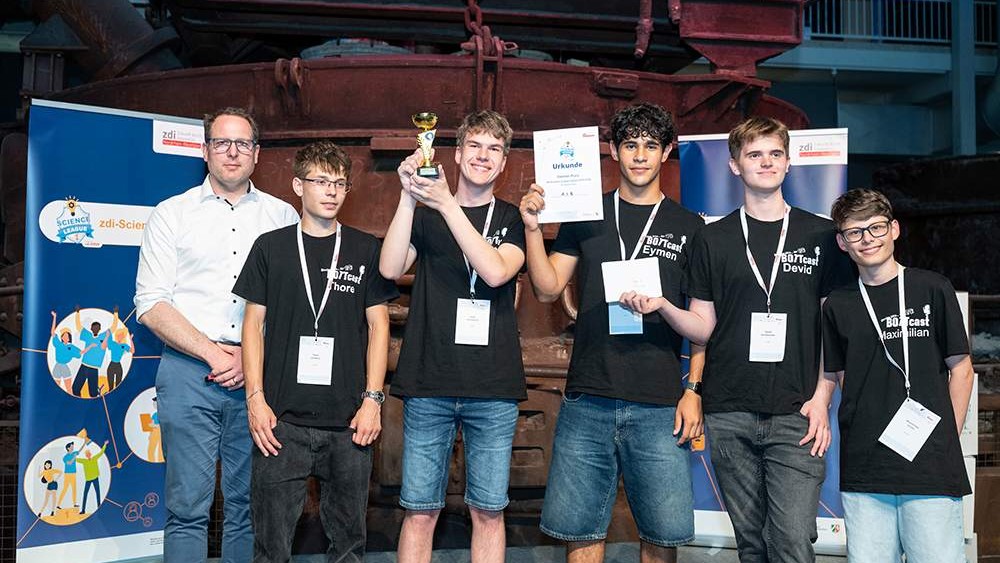 Sechs junge Männer, von denen fünf einheitliche schwarze Hemden mit Namensschildern tragen, stehen neben einem Mann in Weiß. Vor den Bannern halten sie einen Pokal und Urkunden in den Händen.