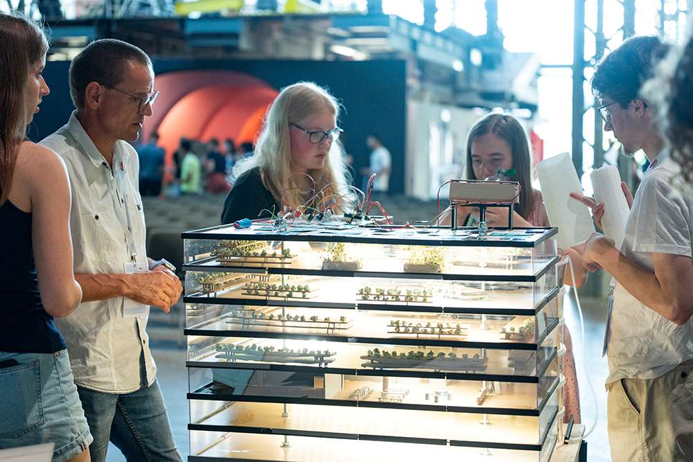 Fünf Personen stehen um ein beleuchtetes, mehrstöckiges Architekturmodell herum. Sie betrachten dessen Details und unterhalten sich in einem Ausstellungsraum.