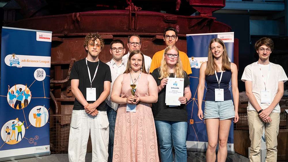 Acht junge Erwachsene stehen in einem Raum und posieren gemeinsam bei einem Wissenschaftswettbewerb, wobei sie eine Trophäe und Urkunden in den Händen halten.