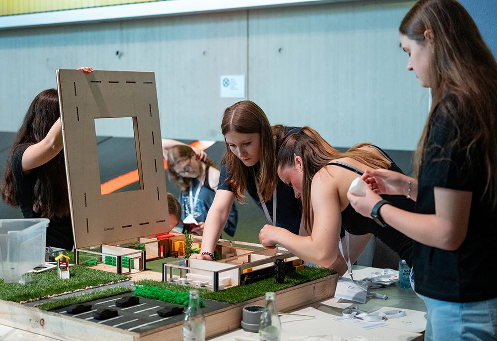 Vier junge Frauen sitzen gemeinsam an einem Tisch und bauen aus verschiedenen Materialien ein Architekturmodell im Miniaturformat zusammen.
