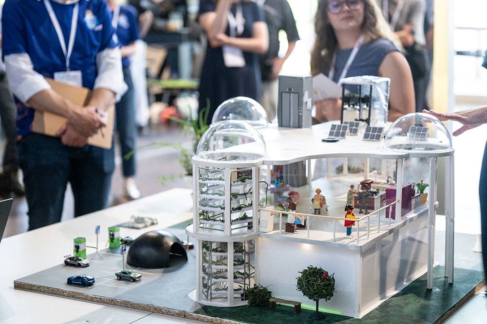 Eine Gruppe von Menschen steht um ein detailreiches Architekturmodell herum, auf dem Miniaturfiguren, Solarmodule, Pflanzen und Elektroautos zu sehen sind.