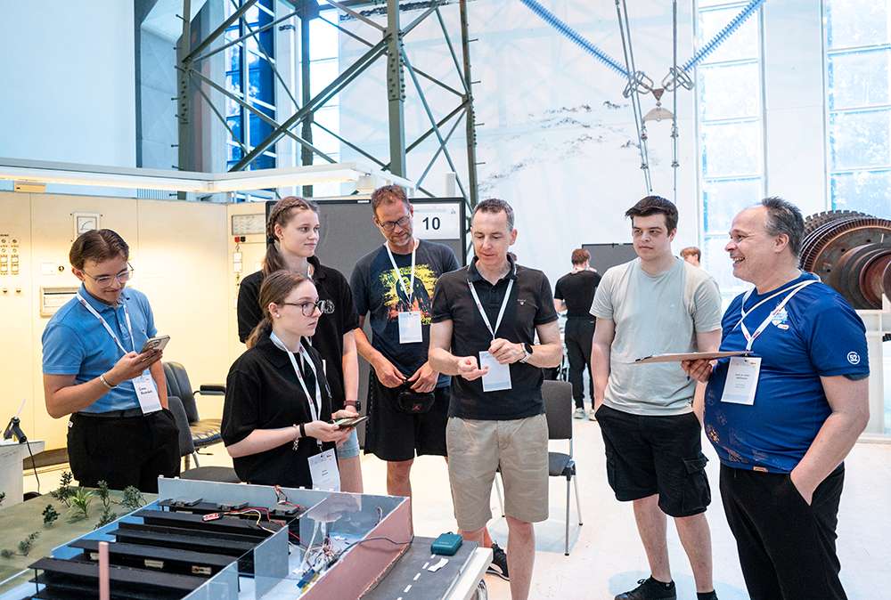 Sechs Personen stehen bei einer Veranstaltung in einem Raum um einen Tisch mit elektronischen Geräten herum und unterhalten sich. Eine weitere Person macht sich auf einem Klemmbrett Notizen.
