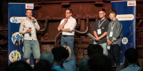 Vier Männer stehen auf einer Bühne vor einem Publikum. Auf zwei Transparenten steht „zdi-Science League“. Ein Mann spricht in ein Mikrofon.