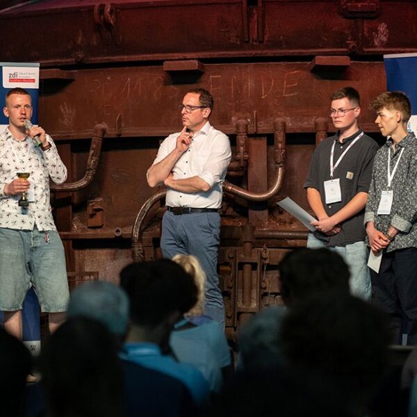 Vier Männer stehen auf einer Bühne vor einem Publikum. Auf zwei Transparenten steht „zdi-Science League“. Ein Mann spricht in ein Mikrofon.