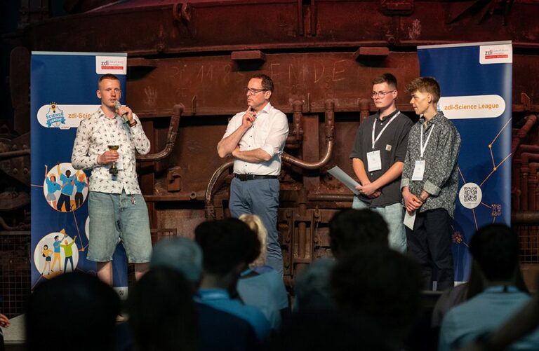 Vier Männer stehen auf einer Bühne vor einem Publikum. Auf zwei Transparenten steht „zdi-Science League“. Ein Mann spricht in ein Mikrofon.