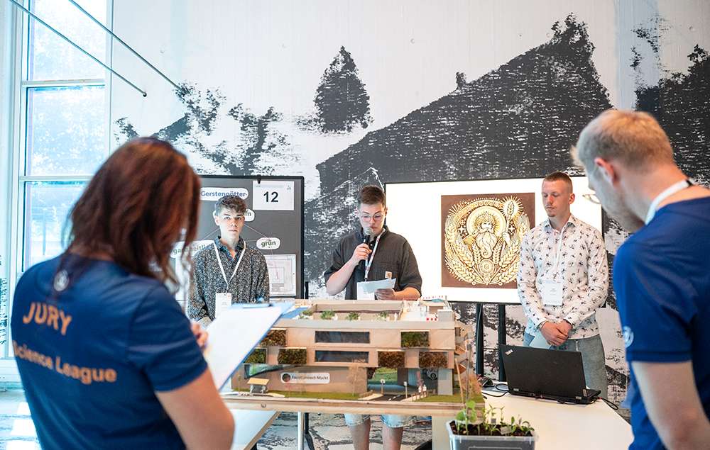 In einem hell beleuchteten Raum, an dessen Wand ein Wandgemälde zu sehen ist, präsentieren die Teilnehmer einer Jury ein Architekturmodell.