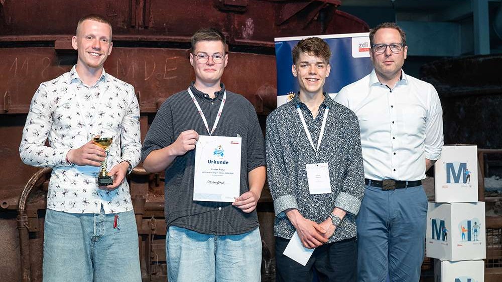 Vier Männer stehen in einer Reihe. Ein Mann hält einen Pokal in der Hand, ein anderer eine Urkunde. Bei dieser Veranstaltung in der Halle tragen sie Namensschilder.