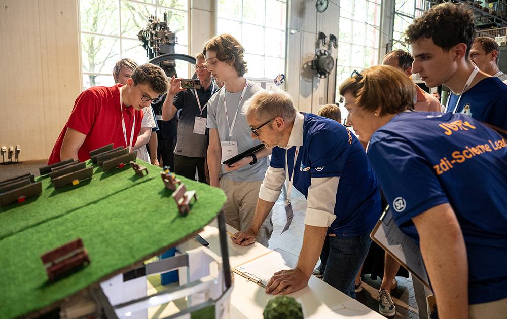 Mehrere Personen versammeln sich um einen Tisch, auf dem ein grünes Modell steht, und eine Person beugt sich vor, um etwas auf ein Blatt Papier zu schreiben.