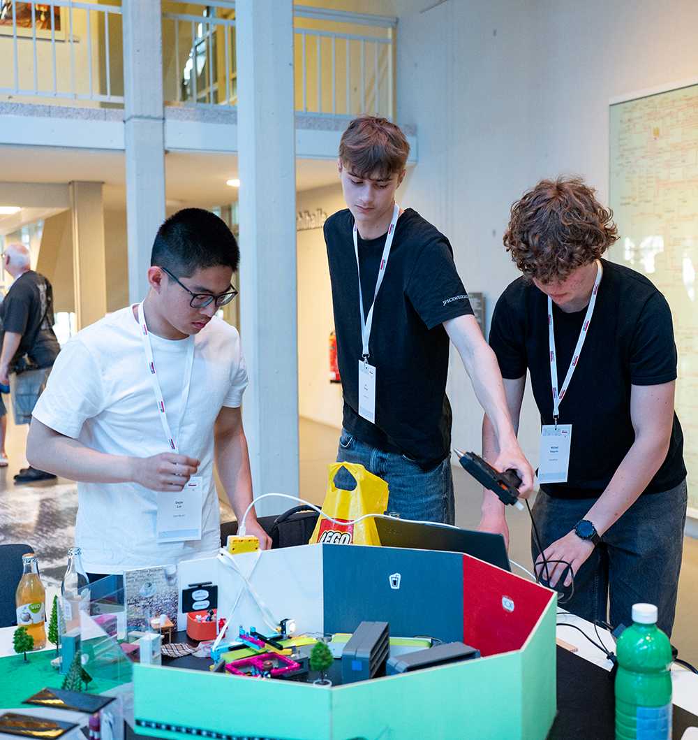 Drei junge Männer mit Namensschildern arbeiten an einem Tisch, auf dem Elektronikgeräte, Kabel, eine LEGO-Tüte, Getränke und andere Gegenstände liegen.