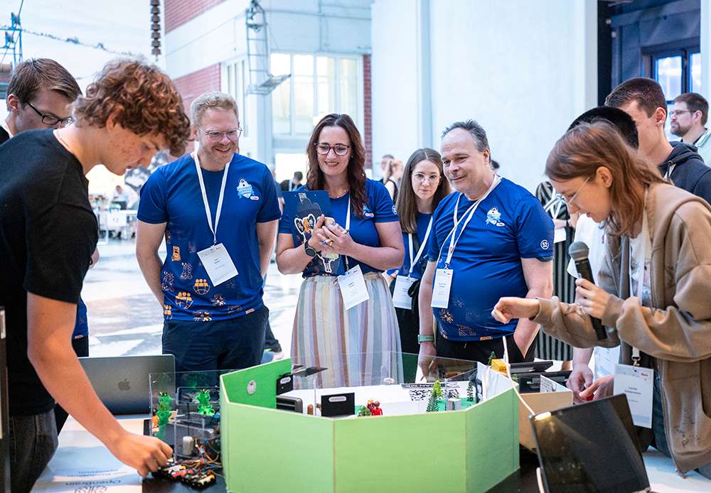 Eine Gruppe von Menschen hat sich um einen Tisch mit Robotikausrüstung versammelt. Einige tragen blaue Hemden und Namensschilder. Einer spricht in ein Mikrofon.