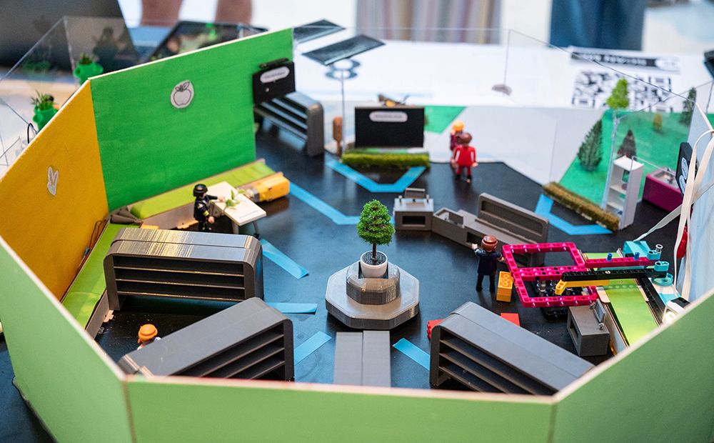 Das Bild zeigt ein Tischmodell mit Miniaturfiguren, Bäumen und Möbeln in einer farbenfrohen, achteckigen Anordnung, die eine Innenraumszene darstellt.