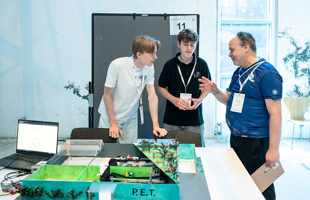 Drei Personen stehen um einen Tisch herum, auf dem ein Robotikprojekt mit der Aufschrift „P.E.T.“ und ein Laptop liegen, und besprechen das Projekt.