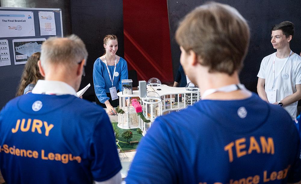 Mehrere Personen stehen um einen Tisch herum, auf dem ein Modell ausgestellt ist. Zwei von ihnen tragen blaue T-Shirts mit den Aufschriften „Jury“ und „Team“. Die anderen tragen weiße Umhängebänder und betrachten das Ausstellungsstück.