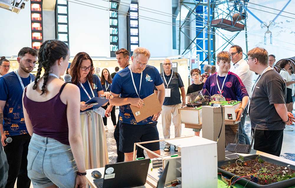 Personen mit Schreibunterlagen beobachten und bewerten Technologieprojekte, die im Rahmen einer Wissenschafts- oder Robotikveranstaltung in einer großen Halle präsentiert werden.