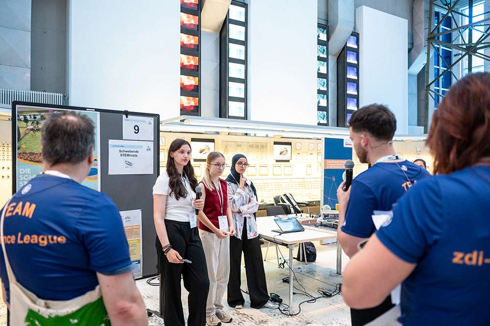 Drei junge Frauen stellen ihr Projekt auf einer Wissenschaftsmesse vor und stehen dabei zwei Männern in blauen Hemden gegenüber. Im Hintergrund sind Plakate und elektronische Geräte zu sehen.