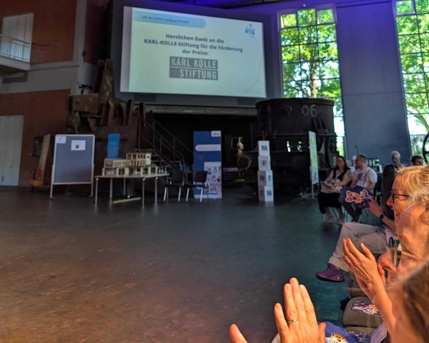 Die Gäste sitzen in einer Halle im Industriestil. Auf einer großen Leinwand wird der Karl-Kölle-Stiftung gedankt, im Vordergrund sind Models zu sehen.