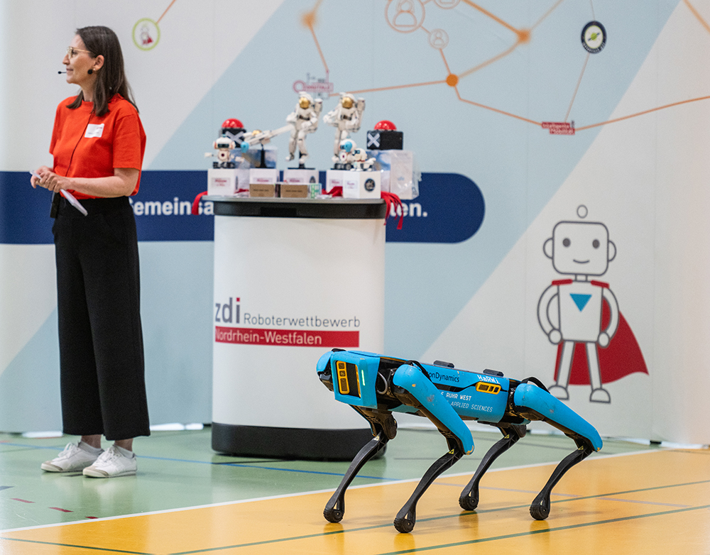Das Foto zeigt einen Roboterhund. Im Hintergrund ist eine Moderatorin zu sehen und ein Tisch mit verschiedenen Pokalen.
