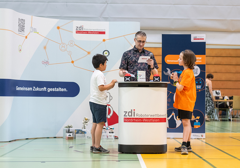 Drei Personen stehen in einer Turnhalle an einem Tisch, auf dem Materialien für einen Robotikwettbewerb liegen. Auf Schildern steht „zdi Roboterwettbewerb Nordrhein-Westfalen“. Zwei Kinder unterhalten sich mit einem Erwachsenen.