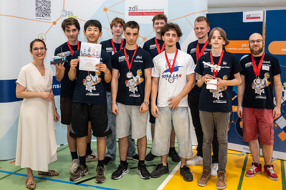 Eine Gruppe junger Menschen, die Medaillen tragen und einheitliche T-Shirts anhaben, steht in einem Raum zusammen mit einer Frau und zwei Männern. Vor den Bannern eines Robotikwettbewerbs halten sie einen Pokal, eine Urkunde und ein Modell in den Händen.