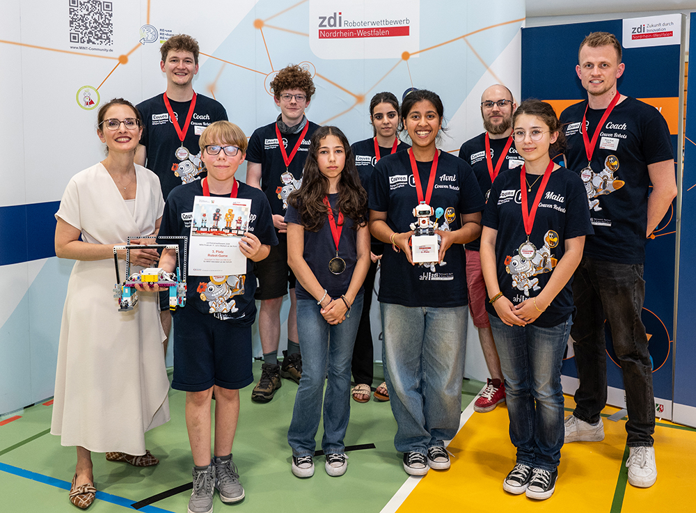 Schüler und Erwachsene befinden sich in einem Raum und posieren bei einem Robotikwettbewerb mit Medaillen, Urkunden und einem Pokal.