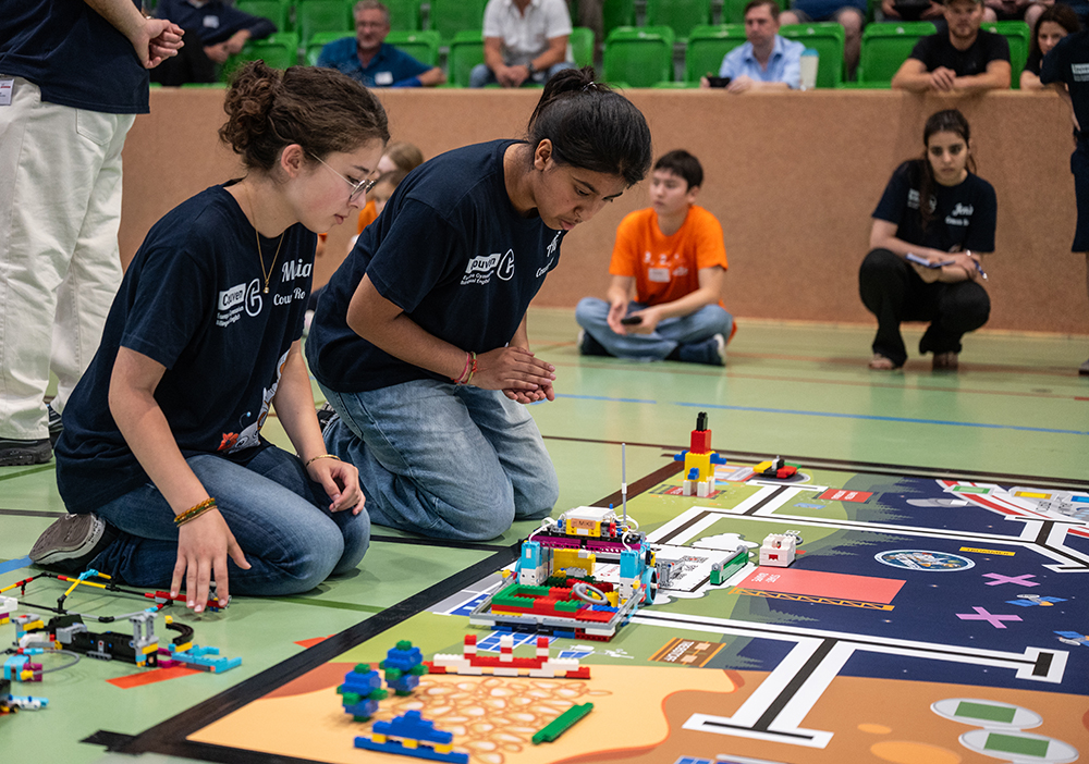 Zwei Schüler knien neben einer Matte für einen Robotikwettbewerb. Sie konzentrieren sich auf LEGO-Modelle und einen Roboter, während andere im Hintergrund in einer Turnhalle zuschauen.