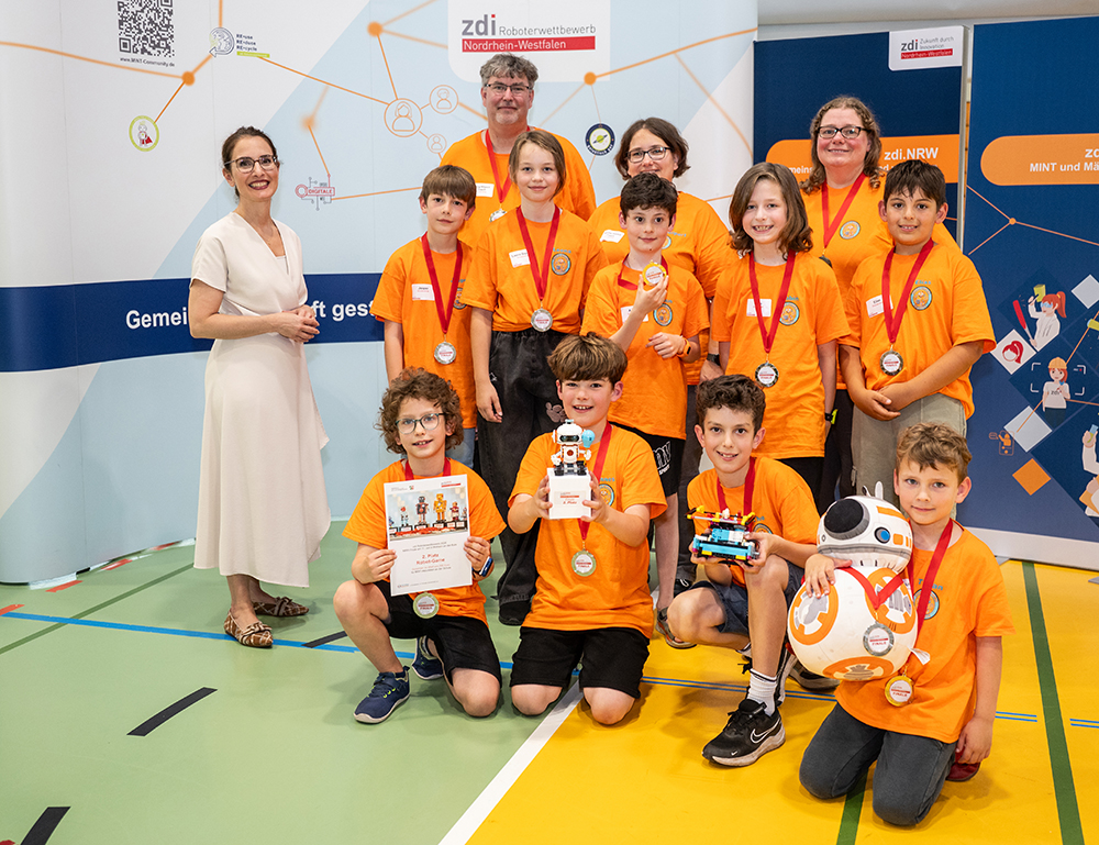 Kinder und Erwachsene in orangefarbenen T-Shirts stehen gemeinsam auf dem Boden einer Turnhalle. Sie halten Medaillen, Urkunden und Roboter in den Händen, während sie vor Bannern mit wissenschaftlichen Motiven in der Halle posieren.