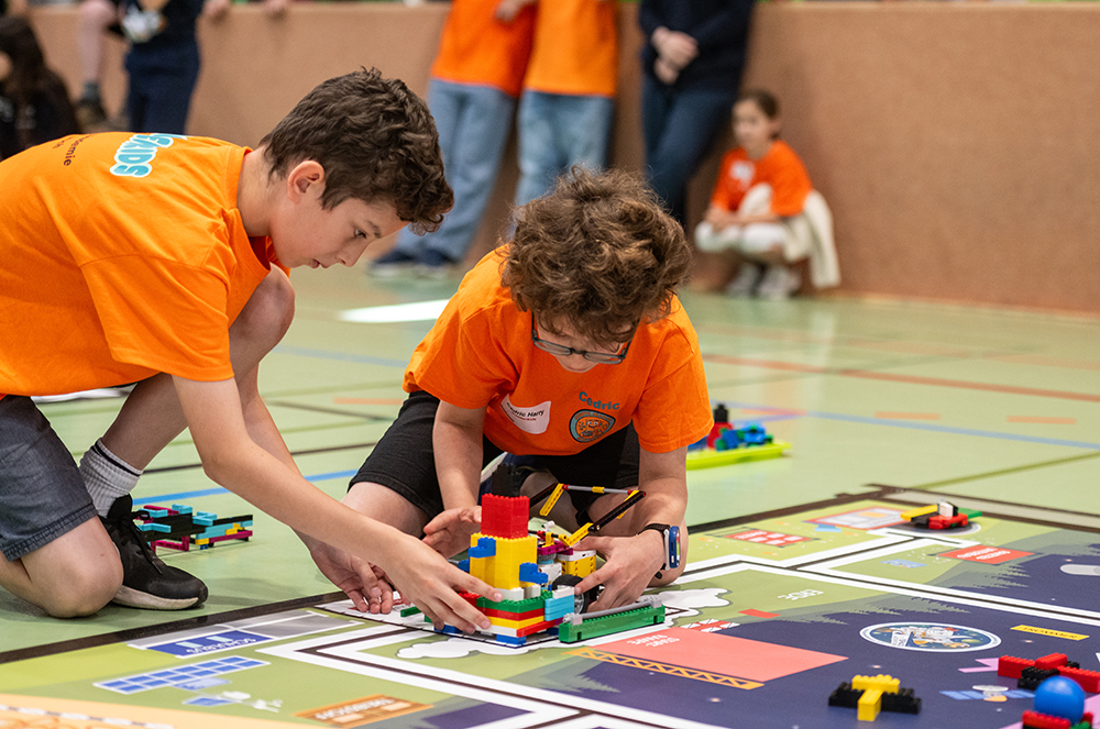 Zwei Kinder tragen orangefarbene T-Shirts und bauen auf einer Matte in einer Turnhalle ein buntes LEGO-Modell zusammen. Im Hintergrund sind weitere Kinder und Erwachsene zu sehen.