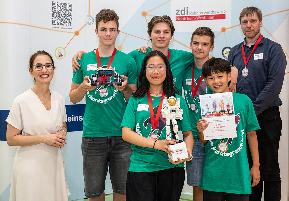Fünf Schüler mit Medaillen und ein Erwachsener posieren bei einem Robotikwettbewerb. Sie stehen neben einem Pokal, einem Roboter und einer Urkunde.