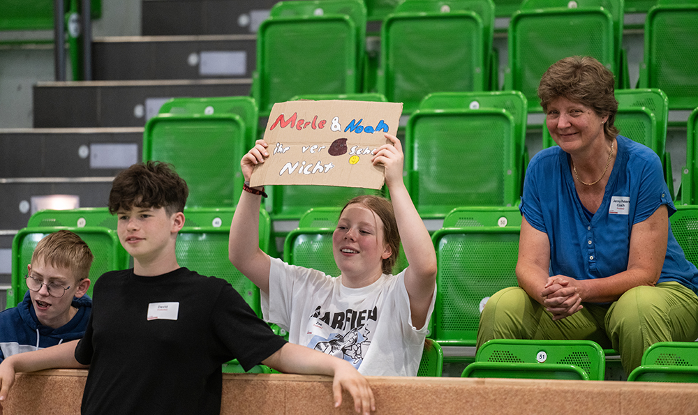 Vier Personen sitzen auf grünen Stadion-Sitzen. Eine Person hält ein selbstgemachtes Schild mit deutschem Text hoch. Sie alle verfolgen eine Veranstaltung.