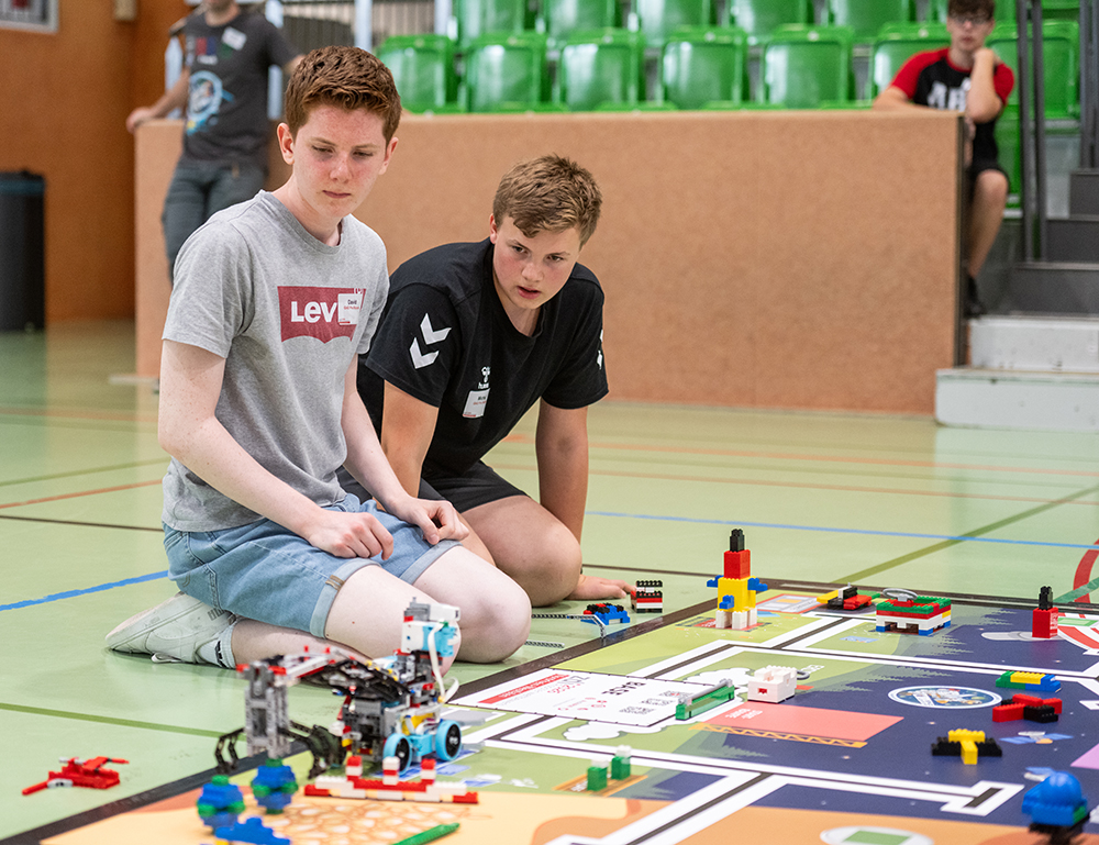 Zwei Jungen knien auf dem Boden einer Turnhalle und beobachten einen LEGO-Roboter, der auf einer Wettkampfmatte steht, umgeben von verschiedenen LEGO-Konstruktionen und -Teilen. Im Hintergrund sind grüne Tribünen und andere Teilnehmer zu sehen.