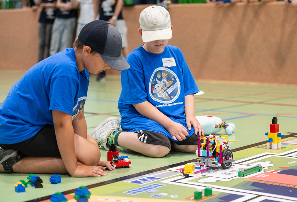 Zwei Kinder in blauen Hemden und Mützen sitzen auf dem Boden einer Turnhalle. Sie sind damit beschäftigt, auf einer Wettkampfmatte LEGO-Konstruktionen zu bauen und anzuordnen.