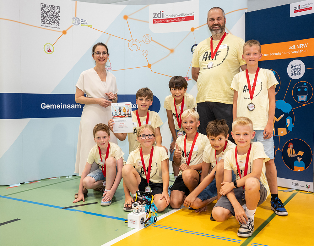 Eine Gruppe von Kindern und zwei Erwachsene befinden sich in einem Raum und posieren mit Medaillen, einer Urkunde und einem kleinen Roboter. Sie stehen vor Bannern des zdi-Roboterwettbewerbs Nordrhein-Westfalen.