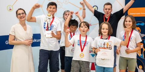 Eine Gruppe von Kindern und zwei Erwachsene stehen bei einem Robotikwettbewerb vor einem Veranstaltungsbanner. Sie feiern mit Medaillen und einem Pokal.