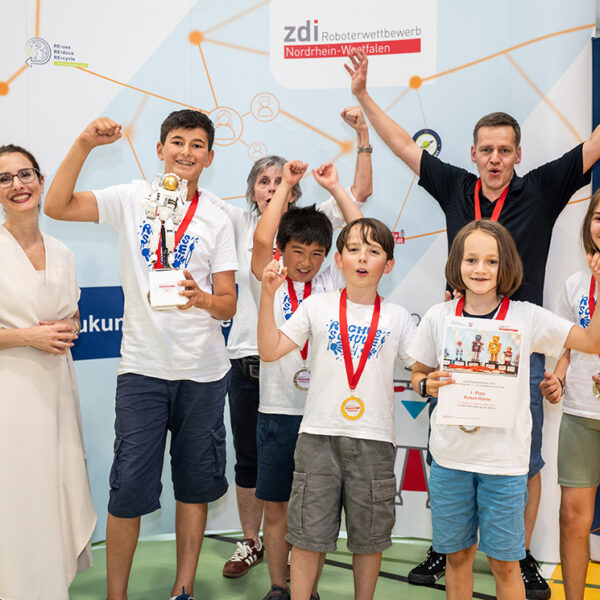 Eine Gruppe von Kindern und zwei Erwachsene stehen bei einem Robotikwettbewerb vor einem Veranstaltungsbanner. Sie feiern mit Medaillen und einem Pokal.