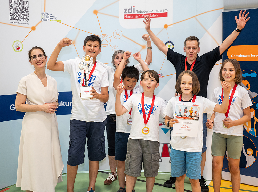 Eine Gruppe von Kindern und zwei Erwachsene stehen bei einem Robotikwettbewerb vor einem Veranstaltungsbanner. Sie feiern mit Medaillen und einem Pokal.
