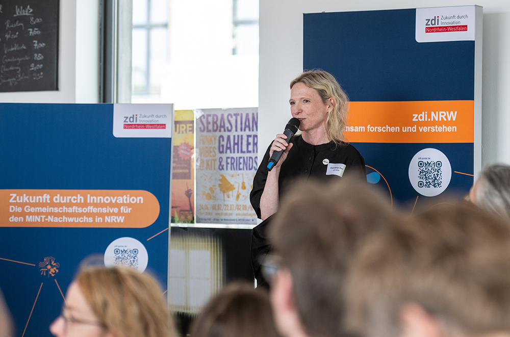 Eine Frau spricht bei einer Veranstaltung in einem Saal, der mit Bannern zum Thema Innovation und MINT in Nordrhein-Westfalen geschmückt ist, in ein Mikrofon. Das Publikum ist zu sehen.